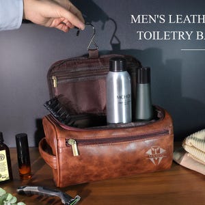 Op de afbeelding: Een bruine leren toilettas met ritssluiting, met verzorgingsproducten. De tas heeft een handvat en een haak om op te hangen. De tekst "MEN'S LEATHER TOILETRY BAG" wordt boven de tas weergegeven.