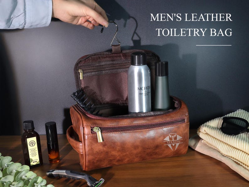 Op de afbeelding: Een bruine leren toilettas met ritssluiting, met verzorgingsproducten. De tas heeft een handvat en een haak om op te hangen. De tekst "MEN'S LEATHER TOILETRY BAG" wordt boven de tas weergegeven.