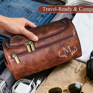 Op de afbeelding: Een bruine leren reistas met gouden ritsen en de letter "M" met geweien. De tas ligt in een koffer met een spijkerbroek, een wit t-shirt, een koptelefoon en een zonnebril. De tekst "Travel-Ready & Compact" staat in de afbeelding.
