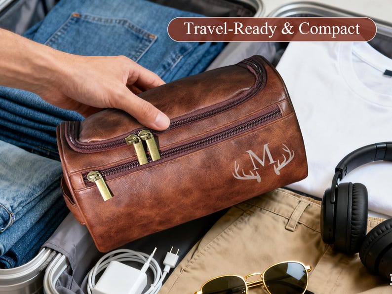 Op de afbeelding: Een bruine leren reistas met gouden ritsen en de letter "M" met geweien. De tas ligt in een koffer met een spijkerbroek, een wit t-shirt, een koptelefoon en een zonnebril. De tekst "Travel-Ready & Compact" staat in de afbeelding.