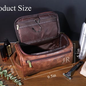 Op de afbeelding: Bruin leren toilettas met ritssluiting, 24 cm lang, 15 cm breed en 13 cm hoog. De tas is open en toont een bruin interieur. De initialen "JR" zijn zichtbaar aan de zijkant. Andere items zijn zichtbaar.