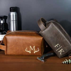 May include: Two brown leather wash bags with personalised initials and a deer antler design. One bag is a lighter brown with the letter "M" and antlers, the other is a darker brown with the initials "F|K".