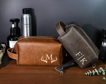 May include: Two brown leather toiletry bags with personalized initials and a deer antler design. One bag is a lighter brown with the letter "M" and antlers, the other is a darker brown with the initials "F|K".