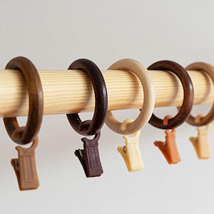 May include: A collection of wooden curtain rings in various shades of brown and beige, each with a matching clip. The rings are attached to a light-colored wooden rod, creating a simple, natural aesthetic.