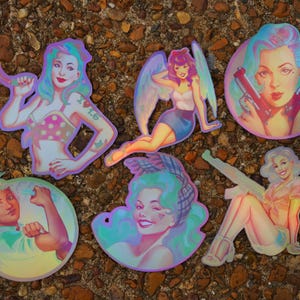 May include: A collection of six holographic stickers featuring pin-up style illustrations. The stickers depict women in various poses, some with wings or holding weapons. The color palette includes pink, blue, and purple hues, with a shimmering, iridescent effect.