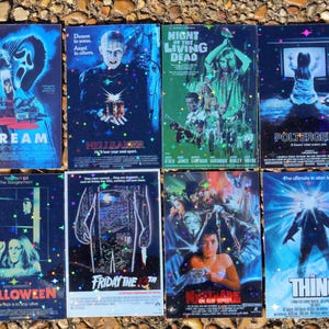 Op de afbeelding: Een verzameling van acht vintage horrorfilm VHS-tapes. Titels zijn onder meer "Scream", "Hellraiser", "Night of the Living Dead", "Poltergeist", "Halloween", "Friday the 13th", "Nightmare on Elm Street" en "The Thing". De tapes liggen op een getextureerd oppervlak.