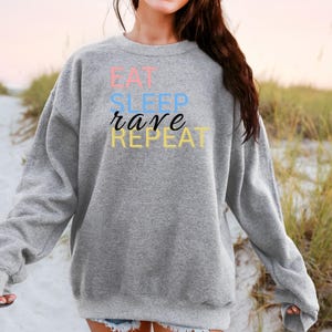 Könnte beinhalten: Ein hellgraues Sweatshirt mit dem Aufdruck "EAT SLEEP rave REPEAT" in Pastellfarben. Das Sweatshirt hat lange Ärmel und einen Rundhalsausschnitt.