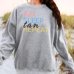 Könnte beinhalten: Ein graues Sweatshirt mit dem Aufdruck "EAT SLEEP tan REPEAT" in Rosa, Blau, Schwarz und Gelb. Das lässige Sweatshirt ist ideal für den Alltag und hat eine entspannte Passform.