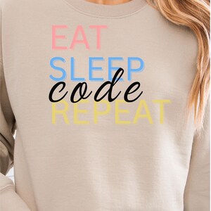 Könnte beinhalten: Beiges Sweatshirt mit dem Text "EAT SLEEP CODE REPEAT" in Rosa, Blau, Schwarz und Gelb. Das Sweatshirt hat eine neutrale Farbe und lange Ärmel. Der Text ist vertikal gestapelt.