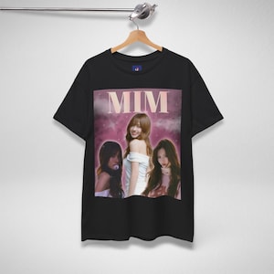 May include: Black t-shirt with a graphic of three women and the text "MIM" in pink. The image has a pink and purple background. The t-shirt is displayed on a wooden hanger.