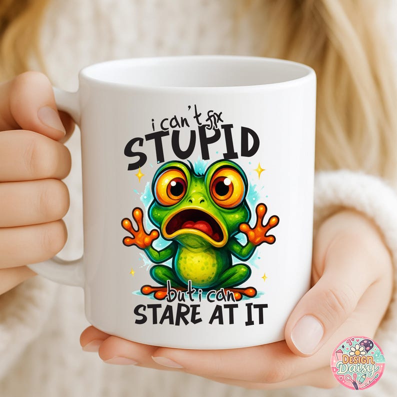 May include: White ceramic mug featuring a cartoon frog with large, expressive eyes and the text "I can't fix STUPID but I can STARE AT IT." The frog is green with orange accents. The mug is held by a person.