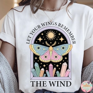 May include: Cream-colored t-shirt featuring a colorful moth design within a framed arch. The moth has blue, pink, and yellow wings. The text "LET YOUR WINGS REMEMBER THE WIND" is arched above and below the moth illustration. Crystals are at the bottom.