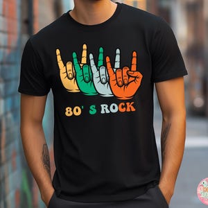 May include: Black t-shirt featuring a colorful graphic of four hands making the rock and roll hand gesture. The hands are in orange, green, light blue, and orange. The text "80's ROCK" is below the hands in orange, green, and blue.
