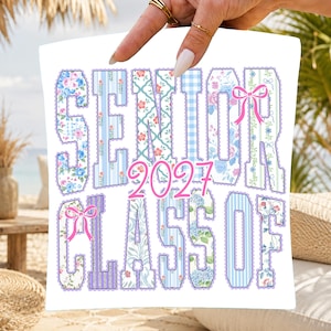 Op de afbeelding: Wit vierkant kussen met de woorden "SENIOR CLASS OF 2027" in een bloemen- en patroonontwerp. De tekst is in verschillende tinten blauw, roze en paars, met roze strikken. Het kussen wordt door een hand vastgehouden.