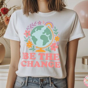 May include: White t-shirt with a colorful graphic of a globe surrounded by flowers and a rainbow. The text "BE THE CHANGE" is printed in pink. The shirt is a crew neck style.