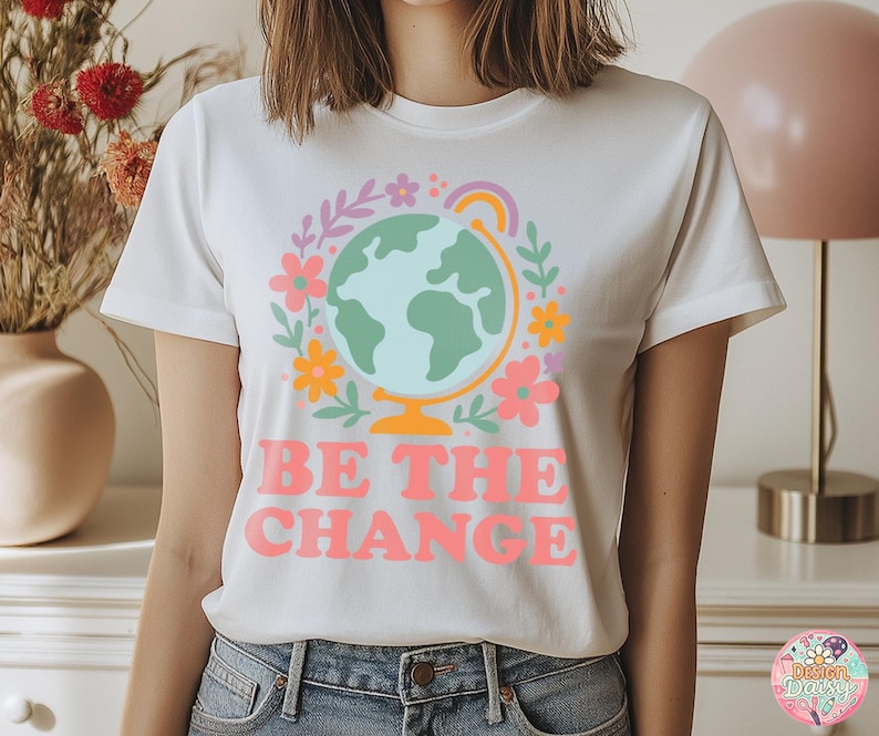 May include: White t-shirt with a colorful graphic of a globe surrounded by flowers and a rainbow. The text "BE THE CHANGE" is printed in pink. The shirt is a crew neck style.