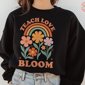 May include: Black sweatshirt with a colorful floral design and the words "TEACH LOVE BLOOM" in orange. The design includes a rainbow and several flowers in shades of orange, pink, and purple. The sweatshirt is a casual style.