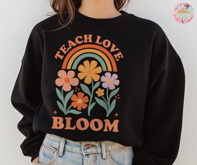 May include: Black sweatshirt with a colorful floral design and the words "TEACH LOVE BLOOM" in orange. The design includes a rainbow and several flowers in shades of orange, pink, and purple. The sweatshirt is a casual style.