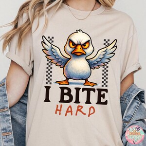 May include: Beige t-shirt featuring a cartoon duck with an angry expression, wings spread, and the text "I BITE HARD" in black and red. The duck has white feathers, an orange beak and feet, and yellow eyes. Checkered patterns flank the duck.