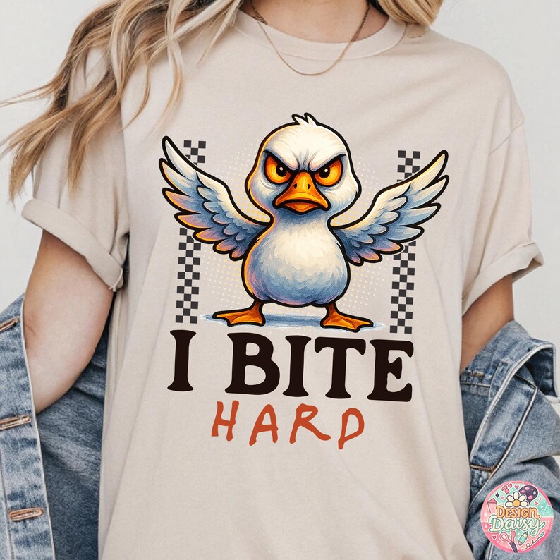 May include: Beige t-shirt featuring a cartoon duck with an angry expression, wings spread, and the text "I BITE HARD" in black and red. The duck has white feathers, an orange beak and feet, and yellow eyes. Checkered patterns flank the duck.