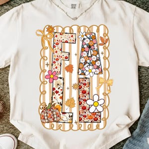 May include: Cream-coloured t-shirt featuring the word "FALL" in a decorative design. The letters are filled with floral and pumpkin patterns, surrounded by autumn-themed graphics like leaves and bows. The design is framed by a scalloped border.