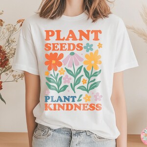 May include: White t-shirt with a colorful floral design and the text "PLANT SEEDS PLANT KINDNESS" in orange, blue, and pink. The graphic features various flowers in orange, pink, and yellow with green leaves.