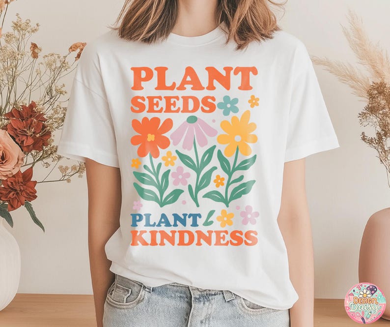 May include: White t-shirt with a colorful floral design and the text "PLANT SEEDS PLANT KINDNESS" in orange, blue, and pink. The graphic features various flowers in orange, pink, and yellow with green leaves.