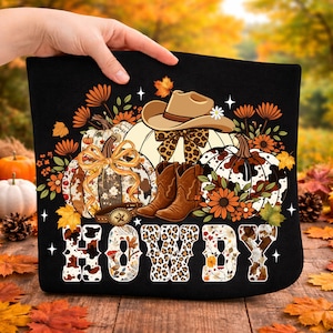 Könnte beinhalten: Schwarze Tragetasche mit einem herbstlichen Design. Das Design umfasst Kürbisse, Cowboy-Stiefel, einen Cowboyhut und das Wort "HOWDY" in einer dekorativen Schriftart. Die Kürbisse haben Blumen- und Kuhfellmuster mit orangefarbenen und braunen Akzenten.
