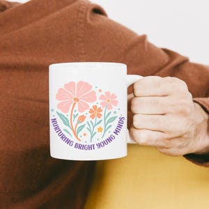 May include: White ceramic mug featuring a floral design in pink, orange, and teal, with the text "Nurturing Bright Young Minds" in a circular layout. The mug has a white handle and is held by a person.