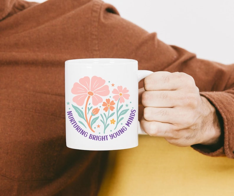 May include: White ceramic mug featuring a floral design in pink, orange, and teal, with the text "Nurturing Bright Young Minds" in a circular layout. The mug has a white handle and is held by a person.