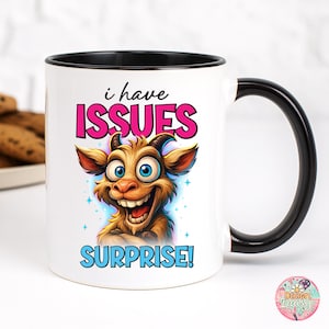 May include: White ceramic mug with a black handle and rim. The mug features a cartoon goat with the text "i have ISSUES SURPRISE!" in pink and blue. The goat has large blue eyes and a wide grin.