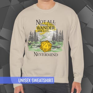 May include: Beige unisex sweatshirt with the text "NOT ALL THOSE WHO WANDER ARE LOS... NEVERMIND." The design includes a yellow D20 die and a mountain and forest scene. The words "UNISEX SWEATSHIRT" are at the bottom.