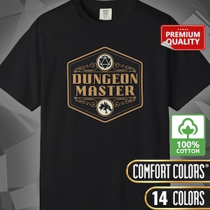 May include: Black t-shirt with a gold and white "Dungeon Master" graphic. The design includes a die, a dragon, and decorative flourishes. The shirt also features a "Premium Quality" tag and a "100% Cotton" label.