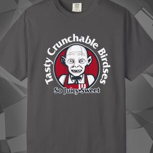 May include: Dark grey t-shirt with a cartoon character inside a red circle. The text reads "Tasty Crunchable Birdses" and "So Juicy-Sweet". The character has pale skin, large eyes, and a toothy grin. The shirt is made of a soft material.