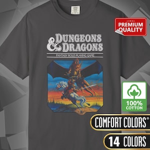May include: Dark grey t-shirt with the "Dungeons & Dragons" logo and fantasy artwork. The shirt includes the text "Fantasy Role-Playing Game" and is made of 100% cotton. Additional text includes "Comfort Colors" and "14 Colors".