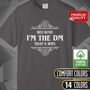 May include: A gray t-shirt with the text "BECAUSE I'M THE DM THAT'S WHY." in white. The shirt has a graphic design with geometric shapes. The shirt is made of 100% cotton and is available in 14 colors.