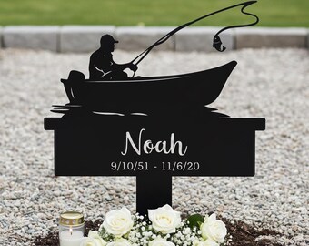 May include: Black metal memorial marker featuring a silhouette of a person fishing from a boat. The name "Noah" and dates 9/10/51 - 11/6/20 are inscribed. A small bouquet of white roses and a candle are at the base.