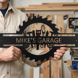 Może przedstawiać: Czarny metalowy szyld garażowy z wyciętym napisem "MIKE'S GARAGE". Znak ma kształt brzeszczotu piły z sylwetkami narzędzi. Znak jest trzymany przez osobę.