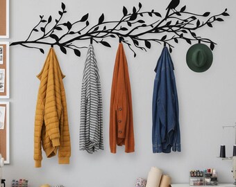 Puede incluir: Un perchero de pared de metal negro con forma de rama de árbol con una silueta de pájaro. Colgados de los ganchos hay una chaqueta acolchada de color amarillo mostaza, una camisa a rayas, una rebeca naranja y una chaqueta vaquera. Un sombrero verde cuelga a la derecha.