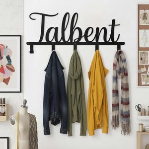 May include: A black metal wall-mounted coat rack featuring the name Talbent in a cursive script font. The rack includes five downward-facing hooks, currently holding a dark blue denim jacket, an olive green hoodie, a yellow jacket, and a scarf.