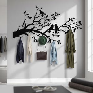 May include: A black metal wall-mounted coat rack in the shape of a tree branch with two birds perched on it. Various coats, a hat, and a purse hang from the hooks. The coats are navy blue, cream, denim, and olive green.