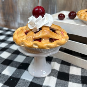 May include: A miniature cherry pie with a lattice crust, topped with whipped cream and a cherry. The pie sits on a white pedestal, with a black and white checkered tablecloth in the foreground. Additional cherries are in the background.