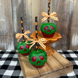 May include: Three decorative poison apple replicas featuring red bases with dripping green skull-shaped icing, topped with black straws patterned with orange pumpkins and finished with orange and white gingham bows on each straw.
