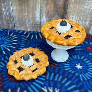 May include: Two miniature blueberry pies with golden-brown lattice crusts, each topped with a dollop of white whipped cream and a single dark blueberry. One pie rests on a small white pedestal stand, while the other sits directly on the surface.