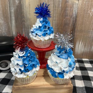 May include: Three decorative cupcakes with blue and white frosting and tinsel toppers. One cupcake sits on a red pedestal, while the others are on a wooden base. The cupcakes have a patriotic theme.