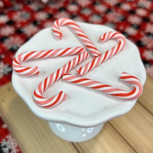 May include: Four candy canes with red and white diagonal stripes arranged on a white ceramic pedestal stand with a scalloped edge. The candy canes are positioned in a circular pattern on the smooth, glossy surface of the white stand.