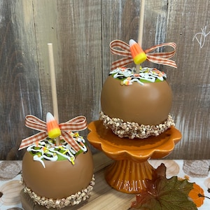 May include: Two caramel-coated apples on wooden sticks, each topped with white icing, colorful sprinkles, and a candy corn piece. The apples feature a brown caramel base with a crunchy nut coating at the bottom and a brown and white gingham ribbon.