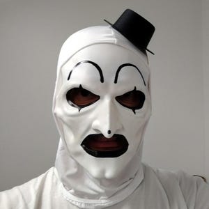 Könnte beinhalten: Eine weiße Clownmaske mit glatter Oberfläche, schwarzen Augenbrauen, schwarzen Augenmarkierungen, einem schwarzen Punkt auf der Nase und einem breiten schwarzen Mund. Die Maske hat eine weiße Kapuze und einen kleinen schwarzen Zylinder obenauf.