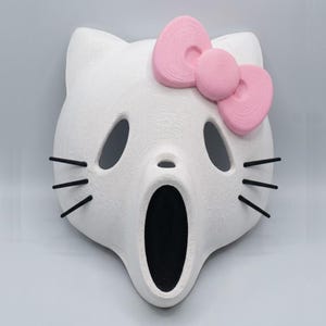 Könnte beinhalten: Eine weiße Maske mit einer katzenartigen Silhouette und einer großen rosa Schleife an der oberen rechten Seite. Die Maske hat zwei ovale Augenöffnungen, ein kleines Nasenloch, eine lange schwarze Mundöffnung und drei dünne schwarze Schnurrhaare.