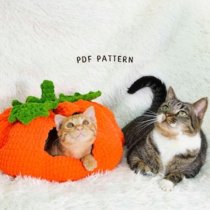 以下が含まれることがあります： 緑の葉が付いたオレンジ色のカボチャ型の編み物猫用ベッドと、中にいるジンジャーキャット。 縞模様の猫がカボチャの隣に座って上を見上げています。「PDF PATTERN」という文字がカボチャの上にあります。